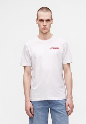 Ung mand med kort hår iført en hvid T-shirt med rødt logo på brystet og blå jeans, stående mod en ensfarvet lys baggrund.