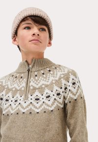 Maglione beige a maglia con colletto alto, chiusura con zip e motivo geometrico bianco. Accompagnato da un berretto a coste rosa chiaro.