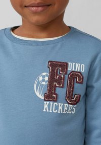 Helles Blaues Sweatshirt mit strukturiertem burgunderfarbenen "FC" und "KICKERS"-Text, einem weißen Fußballgrafik und gestreiftem Halsausschnitt.
