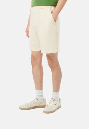 Hombre con pantalones cortos color crema con cintura elástica y bolsillos, calcetas blancas y zapatillas beige sobre un fondo blanco liso.