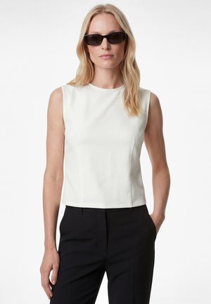 Blondynka w czarnych prostokątnych okularach przeciwsłonecznych, bezrękawniku w białym kolorze i czarnych spodniach, z jedną ręką w kieszeni, na neutralnym tle.
