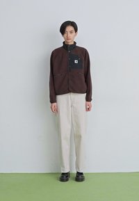 Joven de pie contra una pared lisa, viste una chaqueta de forro polar marrón oscuro con un bolsillo negro, pantalones beige claros y zapatos negros.