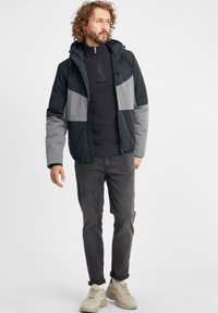 Herrejakke med sorte og grå paneler, glidelås foran, hette og strikket krage. Kombinert med grå jeans og lyse joggesko.