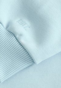 Lyseblå sweatshirtstof med en glat overflade, der har en ribbet manchet og et præget logo-detalje.