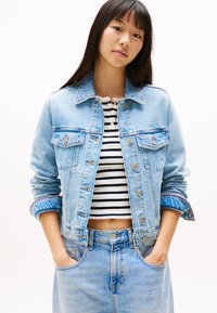 Giacca di jeans azzurro chiaro con chiusure a bottone, tasche sul petto e maniche arrotolate. Indossata su un top corto a righe bianco e nere e una gonna di jeans.