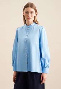 Hellbluse mit hochgeschlossenem Kragen, Knopfverschluss und gepufften langen Ärmeln, aus glattem Stoff gefertigt. Besitzt einen dezenten Glanz und eine taillierte Passform.