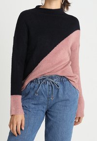 Stickad tröja med en svart och rosa diagonal design, med hög hals och långa ärmar, i kombination med avslappnade jeans med dragsko.