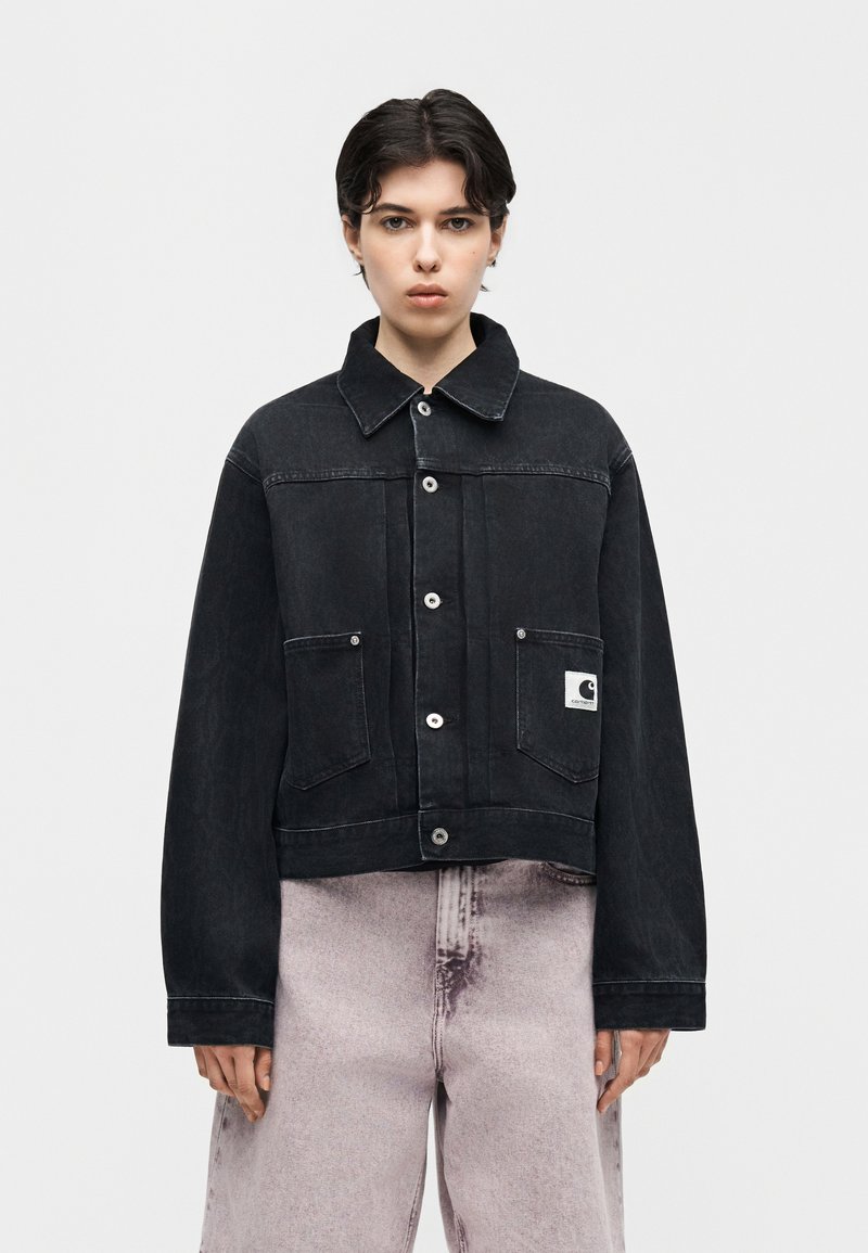 Giovane con capelli corti e scuri indossa una giacca di jeans nera oversize con tasche frontali e jeans larghi a gamba larga dal lavaggio chiaro, in piedi di fronte.