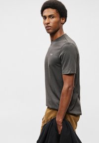 Tumši pelēks kokvilnas T-krekls ar īsām piedurknēm un apaļu kakla izgriezumu, ar nelielu baltu logotipu kreisajā krūtī. Modelis tur tumšu jakas.