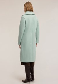 Abrigo verde menta con acabado texturizado, que presenta un cuello ancho, silueta recta y mangas largas, combinado con botas marrones hasta la rodilla.