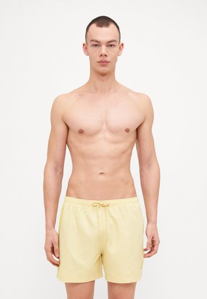 Shorts de bain jaunes à cordon de serrage avec une coupe décontractée, texture lisse et logo subtil sur le côté gauche. Le modèle se tient devant un fond uni.