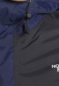 Granatowa i czarna kurtka przeciwdeszczowa o teksturowanej powierzchni, z regulowanym sznurkiem przy szyi oraz białym logo w dolnym prawym rogu.