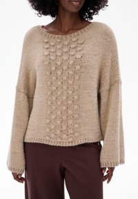 Beżowy sweter dzianinowy z teksturowanym wzorem bąbelków na przodzie, z szerokimi rękawami i luźnym krojem, zestawiony z brązowymi spodniami.