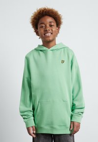 Niño sonriente con cabello rizado, vistiendo una sudadera verde menta con un pequeño logo de un pájaro amarillo y pantalones oscuros, contra un fondo sencillo.