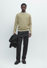 Beige trui, donkere spijkerbroek en zwarte schoenen. Houdt een donker jasje vast. Simpel ontwerp, gestructureerde brei en comfortabele pasvorm.