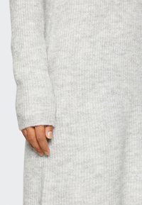 Maglione grigio chiaro a maglia con texture a costine, dotato di maniche lunghe e fori per il pollice. Il tessuto sembra morbido e confortevole, con una vestibilità rilassata.