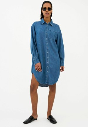 Nő kék farmer ingruhába, fekete napszemüvegbe és fekete slip-on cipőbe öltözve, fehér háttér előtt áll.