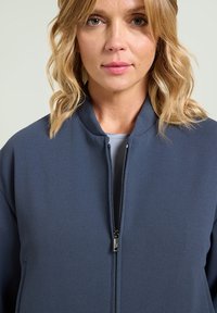 Donkerblauwe bomberjack met een gladde textuur, voorzien van een ritssluiting en een opstaande kraag. Minimalistisch ontwerp zonder zichtbare patronen of accenten.