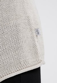 Tessuto lavorato a maglia beige chiaro con una trama sciolta, caratterizzato da una piccola etichetta del marchio sul lato e un sottile dettaglio orlo.