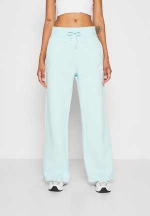 Personne portant un jogging large bleu clair avec taille à cordon et baskets blanches, haut blanc court et tatouages visibles sur les bras.