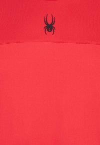 Prenda roja de manga corta hecha de tela suave, con un logo de araña negra en el pecho y un simple detalle de costura horizontal.