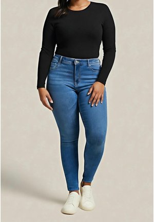 Vrouw draagt een zwarte top met lange mouwen, blauwe high-waisted skinny jeans en witte sneakers, staand tegen een neutrale achtergrond.