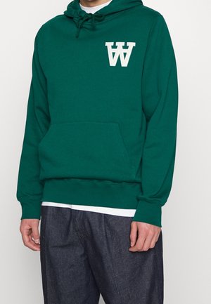 Mężczyzna w ciemnozielonej bluzie z kapturem z białym logo "W" i ciemnoniebieskich dżinsach, stoi na jednolitym tle.