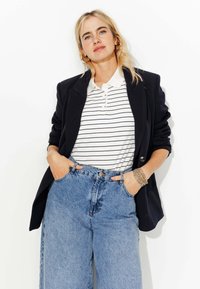Femme blonde portant un blazer noir, un polo à rayures blanches, un jean large taille haute, des boucles d'oreilles en or et des bracelets, les mains dans les poches.