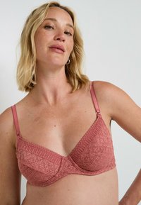 Reggiseno in pizzo di colore rosa tenue con coppe a triangolo, spalline regolabili e dettagli a motivo floreale. Presenta una chiusura a gancio e occhiello.