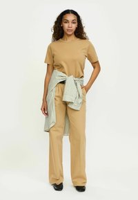 Beidzē bēšas krāsas T-krekls un brīvie bēšas krāsas bikses, ap vidukli sasieta josla ar svītrām gaiši zaļā un baltā krāsā. Melni apavi.