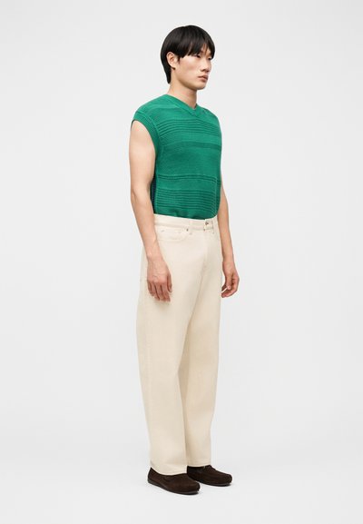 Νέος άνδρας που στέκεται, φορώντας πράσινο πλεκτό αμάνικο γιλέκο, μπεζ παντελόνι με φαρδιά πόδια και σκούρα καφέ παπούτσια, μπροστά σε απλό γκρι φόντο.