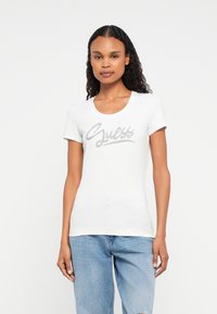Frau mit gewelltem Haar trägt ein weißes enges T-Shirt mit dem silbernen "Guess"-Logo in Schreibschrift und hellblauen, verwaschenen Jeans, stehend vor einem weißen Hintergrund.