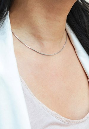 Zilveren metalen ketting, plat ontwerp, dun profiel, gladde textuur, subtiele glans, rustend tegen blote huid. Minimalistische en elegante uitstraling.
