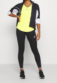Conjunto atlético de Nike que incluye una chaqueta cortavientos negra, una camiseta amarilla brillante y leggings negros con logo. La chaqueta cortavientos tiene detalles en blanco.