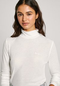 Haut à col roulé blanc côtelé avec des manches longues, présentant une texture subtile et un petit logo brodé sur la poitrine.
