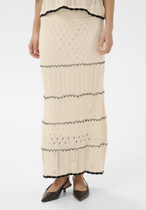 Femme portant une jupe longue tricotée de couleur crème avec des rayures en zigzag noires, associée à des chaussures noires pointues à petit talon.