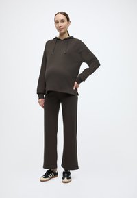 Σκούρο καφέ φούτερ με κουκούλα και παντελόνι φαρδύ, κατασκευασμένα από μαλακό ύφασμα, με άνετη εφαρμογή και μινιμαλιστικό σχεδιασμό. Μαύρα αθλητικά παπούτσια με ρίγες.