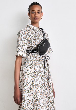 Femme portant une robe blanche à fleurs, un sac bandoulière noir et des boucles d'oreilles créoles dorées, debout devant un fond blanc uni.