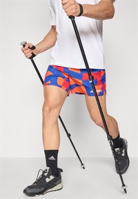 Athletic shorts featuring a multicolored geometric pattern in red and blue, worn with a white t-shirt and black socks, with trekking poles.