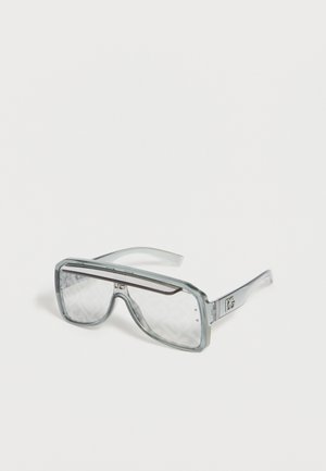 Priehľadné nadmerné obdĺžnikové slnečné okuliare s priehľadným sivým rámom a jemným logom na straniciach, zobrazené na bielom pozadí.