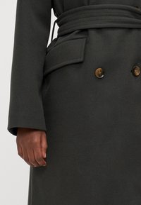 Dobbeltspent mørkegrå frakk med belte, med vide ermer og en lomme. Glatt, myk tekstur med brune knapper som detaljer.