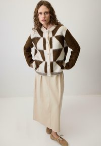 Kobieta z kręconymi włosami, ubrana w kardigan w geometryczny wzór w brązowo-kremowych odcieniach, beżową spódnicę midi i jasnobrązowe baleriny, stoi z rękami na biodrach.