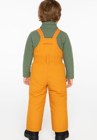 Kind met kort bruin haar, gekleed in een groene trui en oranje sneeuwbibbroek met zwarte laarzen, staande met de rug naar de kijker op een witte achtergrond.