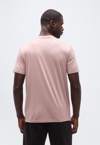 Άνδρας που φοράει απλό ανοιχτό ροζ μπλουζάκι με κοντά μανίκια και μαύρο παντελόνι, όρθιος με την πλάτη του να κοιτάζει προς την κάμερα, μπροστά σε λευκό φόντο.