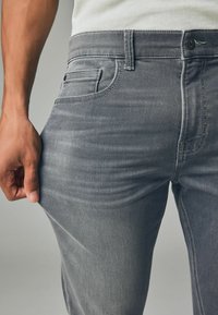 Jeans din denim gri cu un design ajustat, având un buzunar frontal și cusături vizibile. Textura pare ușor uzată cu cute naturale.