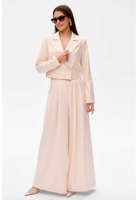 Lichtroze korte blazer met dubbele knopen, gecombineerd met wijde broekspijpen. Accessoires zijn onder andere zwarte zonnebril en gestreepte schoenen.