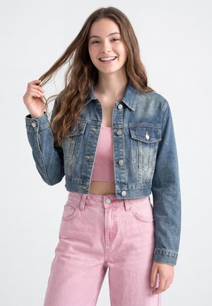Junge Frau lächelt, trägt eine kurze blaue Jeansjacke über einem rosa gerippten Crop-Top und hoch taillierte rosa Jeans, hält eine Haarsträhne.