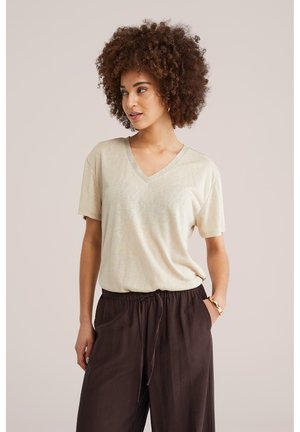 Moteris su garbanotais plaukais, dėvinti smėlio spalvos V formos kaklo marškinėlį ir tamsiai rudus raišteliais suveržtus kelnes, stovinti viena ranka kišenėje.