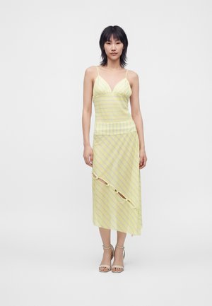 Femme aux cheveux noirs portant une robe jaune rayée sans manches avec une découpe diagonale et des boutons, debout devant un fond blanc uni.