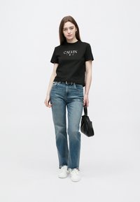 Tânără purtând un tricou negru Calvin, blugi albaștri, adidași albi, ținând o geantă neagră, stând în fața unui fundal alb.